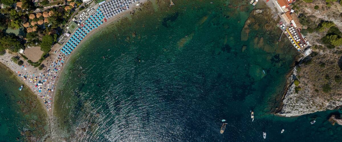 An aerial view of a coastal area with a beach, a swimming pool, and a densely packed row of umbrellas.