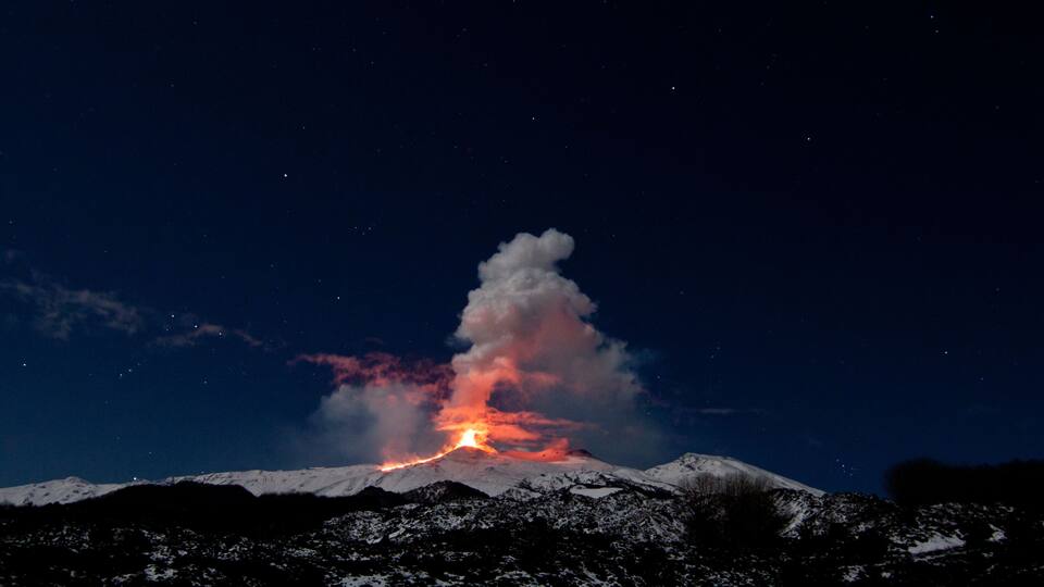 A snow-covered mountain with a volcanic eruption at night.