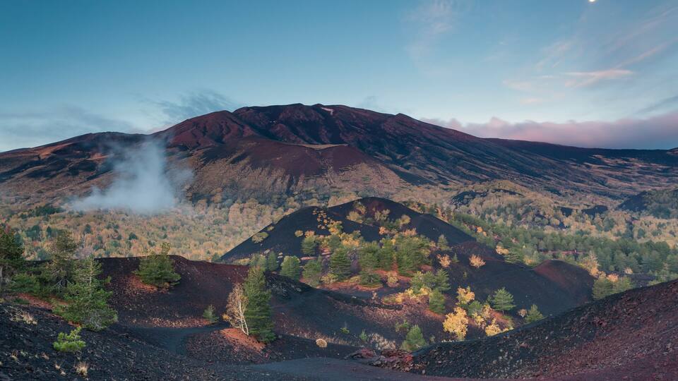 A volcanic landscape with a prominent mountain, a visible smoke plume, and a clear sky with a moon.
