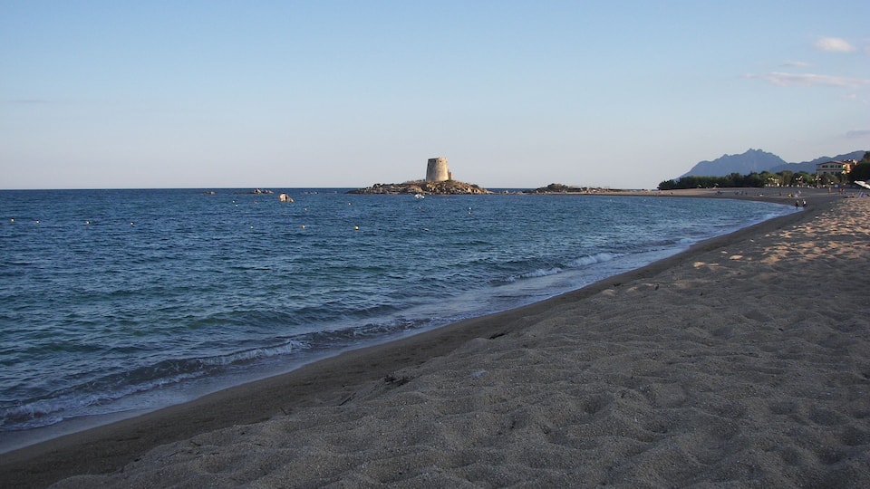 A sandy beach with gentle waves, a rocky outcrop with a tower, and distant mountains under a clear sky.
