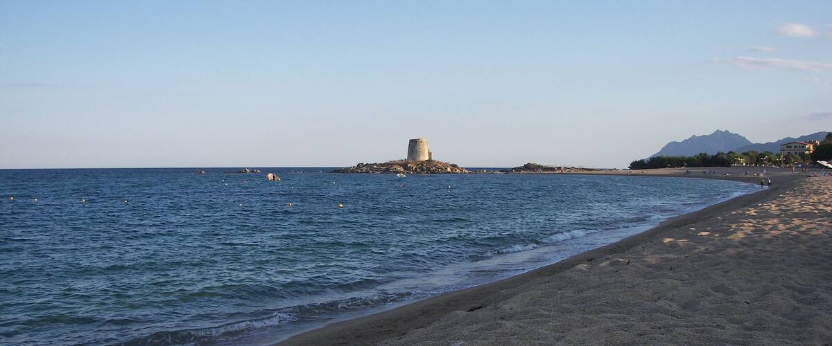 A sandy beach with gentle waves, a rocky outcrop with a tower, and distant mountains under a clear sky.
