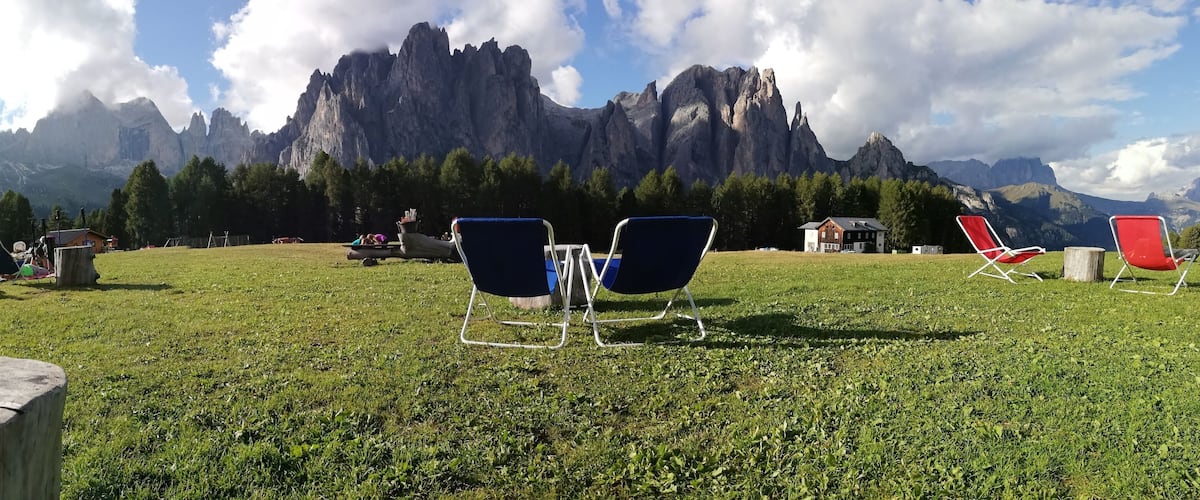 A grassy area with outdoor chairs and a table with a drink, set against a backdrop of mountains.