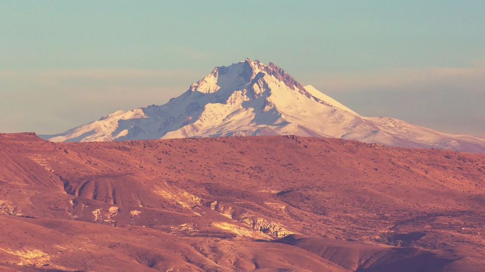A snow-capped mountain with a rugged, reddish-brown landscape in the foreground.