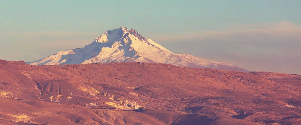 A snow-capped mountain with a rugged, reddish-brown landscape in the foreground.