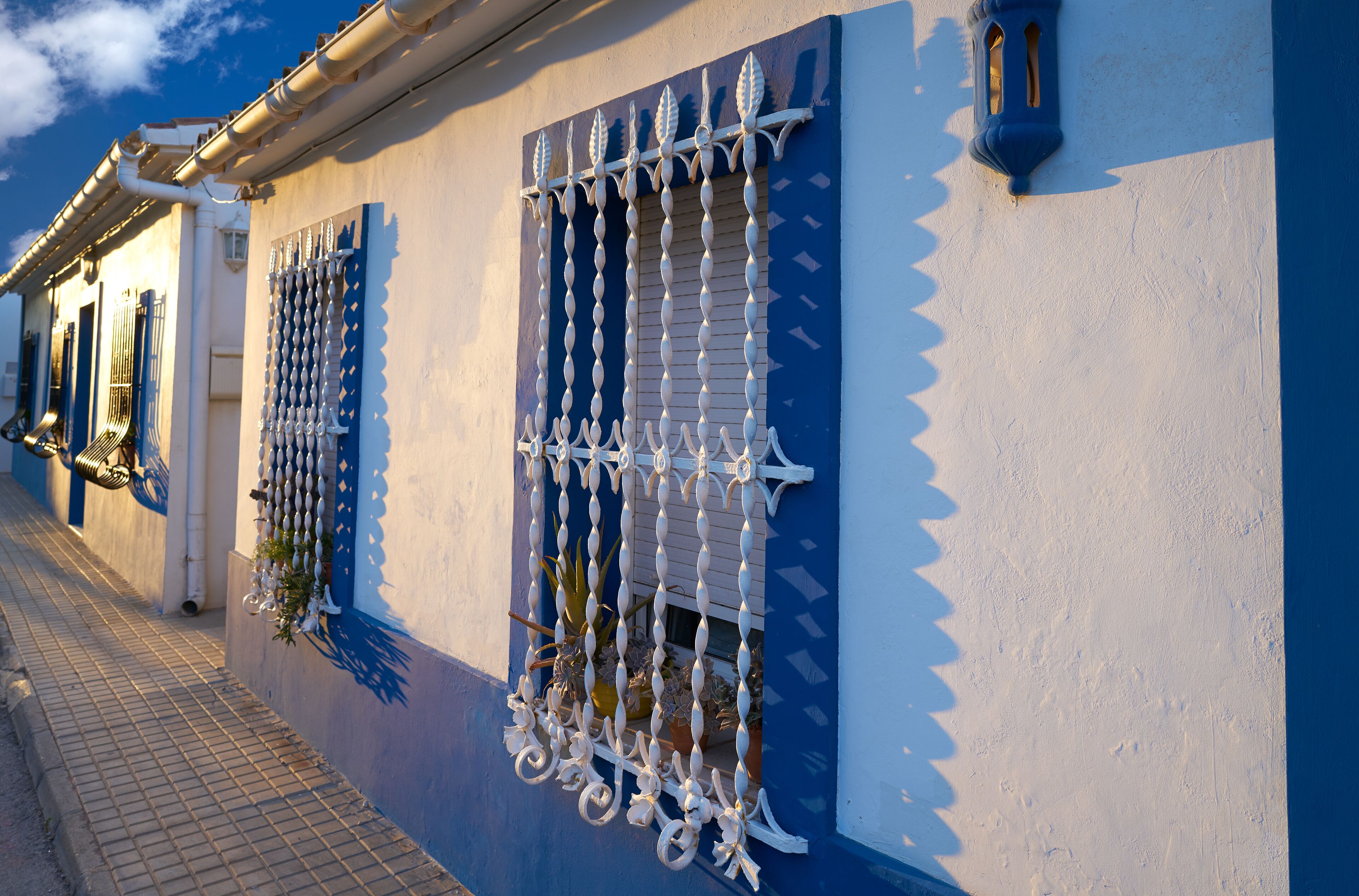 A building with blue and white walls, decorative metal bars on the windows, and a tiled sidewalk.