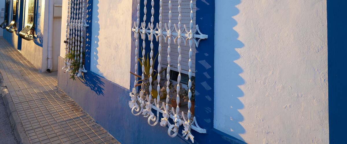 A building with blue and white walls, decorative metal bars on the windows, and a tiled sidewalk.