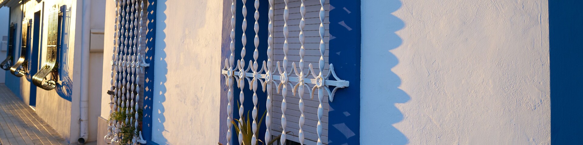 A building with blue and white walls, decorative metal bars on the windows, and a tiled sidewalk.