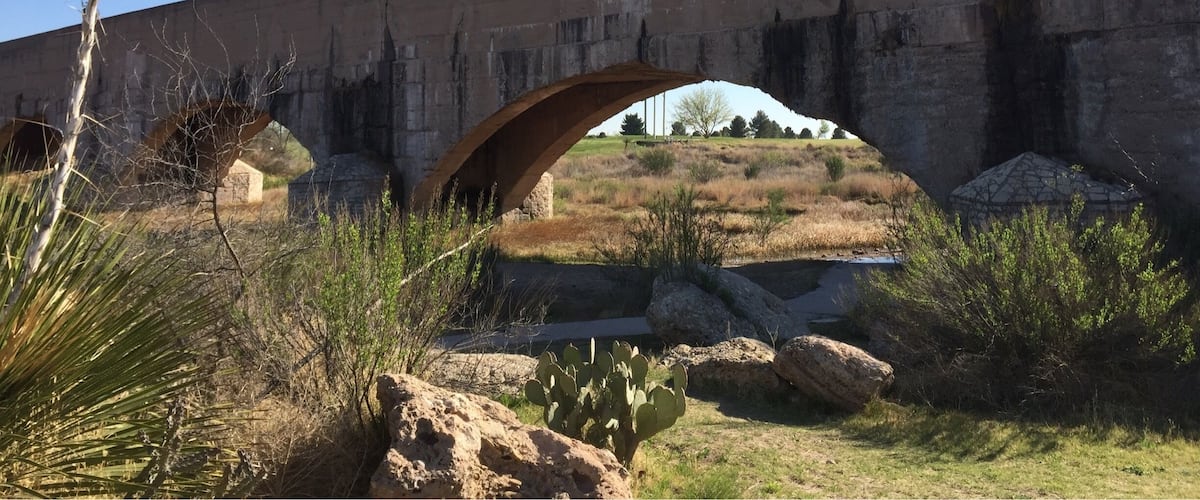 A concrete bridge with an arch over a dry riverbed, surrounded by sparse vegetation and rocks.