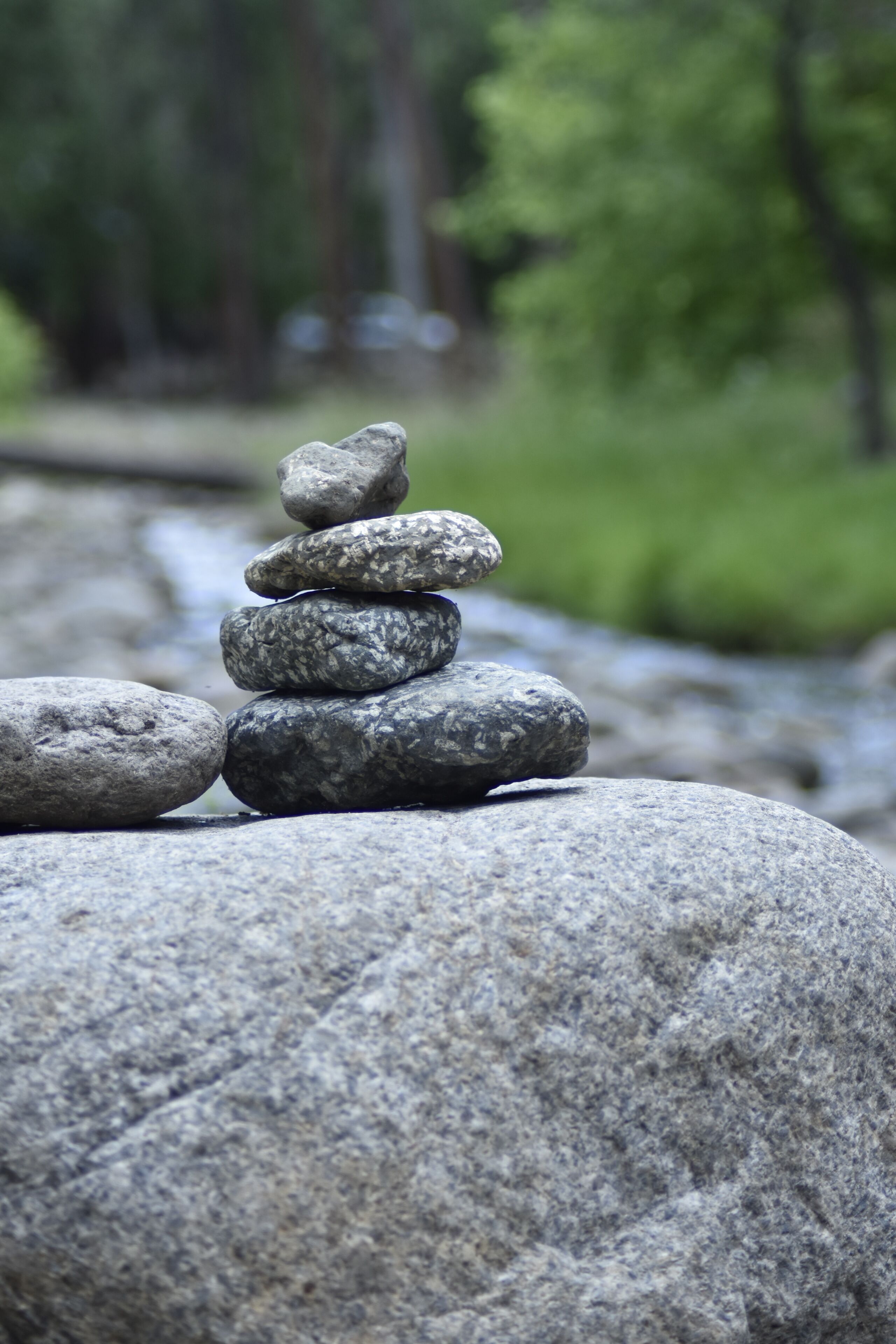 Um conjunto de pedras arredondadas e lisas equilibradas sobre uma rocha maior, com uma floresta e um riacho desfocados ao fundo.
