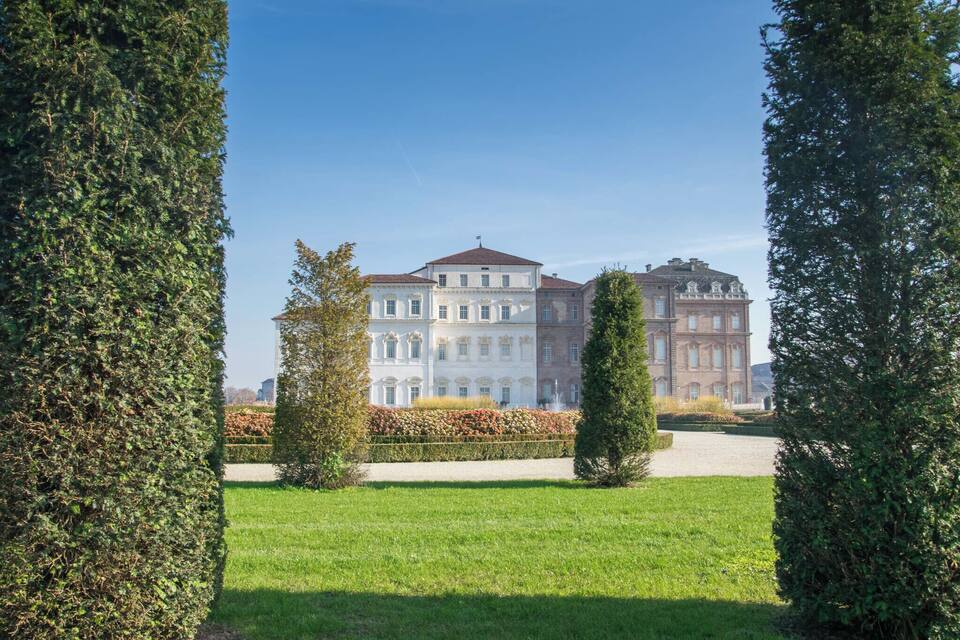 Um edifício grande e clássico, com fachada simétrica, rodeado por jardins bem cuidados e árvores altas e frondosas.