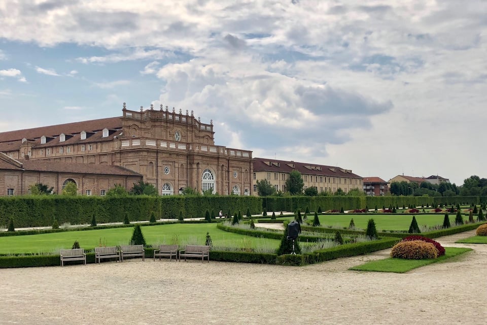 A large historic building with a symmetrical facade, surrounded by well-manicured gardens and hedges.