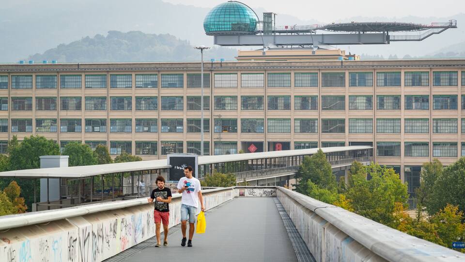 A pedestrian bridge with graffiti, a large building with a dome, and trees in the background.