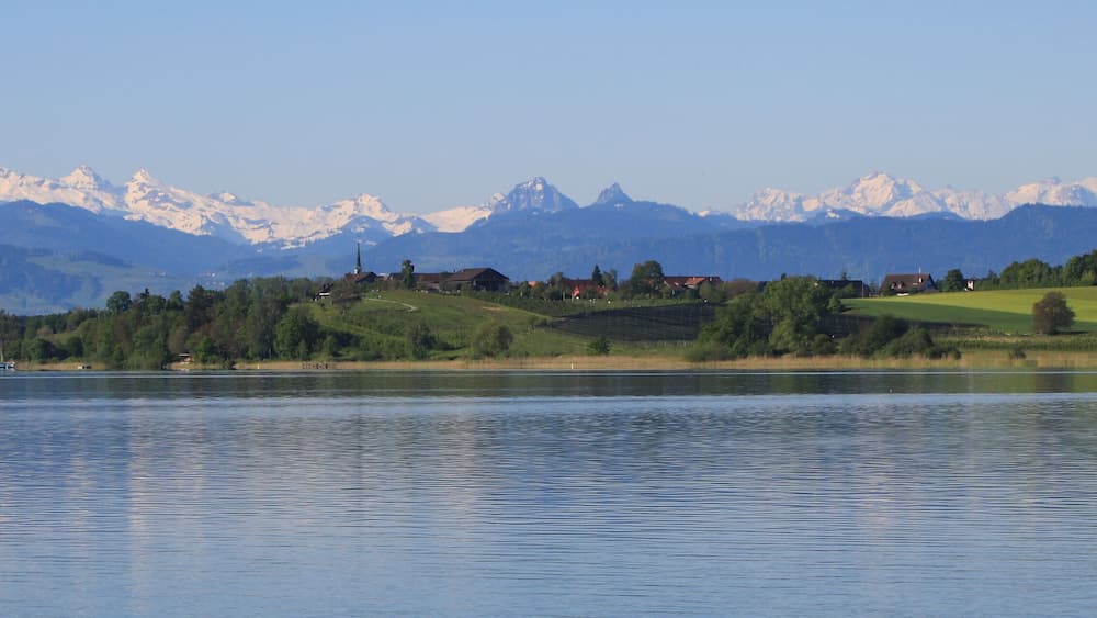 Ein ruhiger See mit einem Boot, umgeben von grünen Hügeln und schneebedeckten Bergen im Hintergrund.