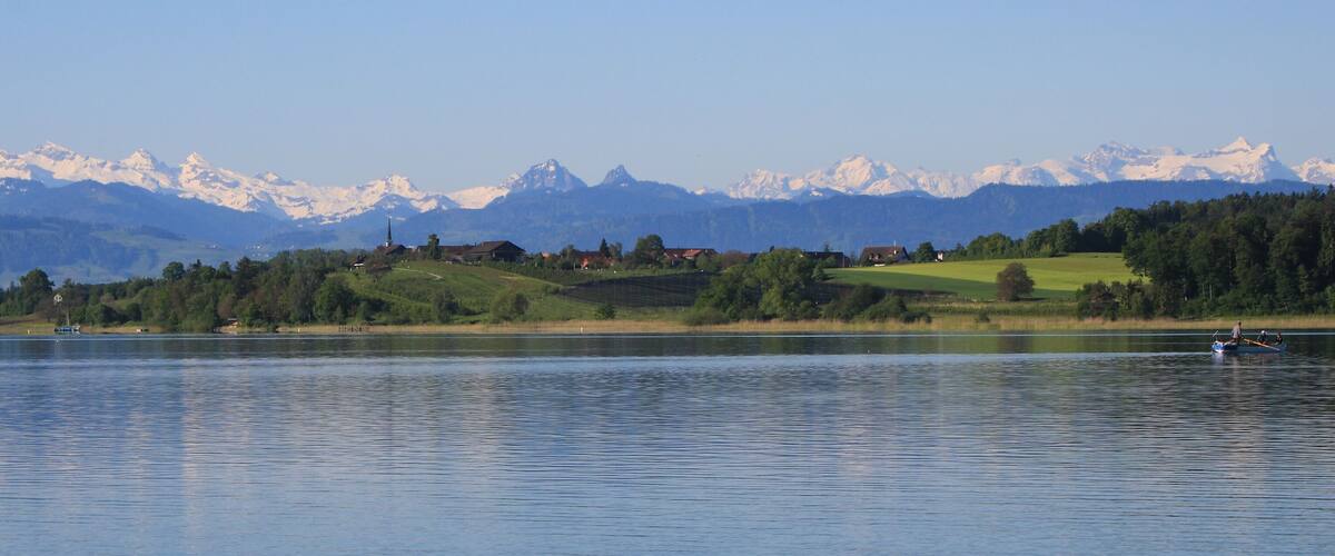 A calm lake with a boat, surrounded by green hills and snow-capped mountains in the background.