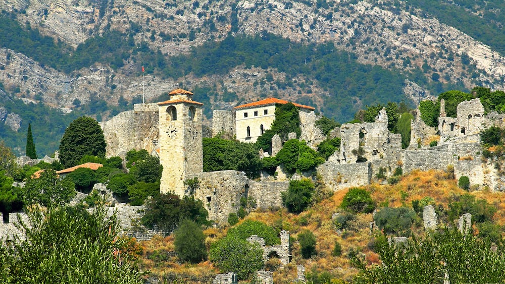 A historic stone fortress with a clock tower and a bell tower, surrounded by trees and rocky terrain.