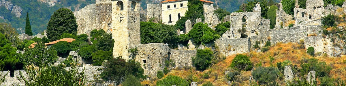 A historic stone fortress with a clock tower and a bell tower, surrounded by trees and rocky terrain.