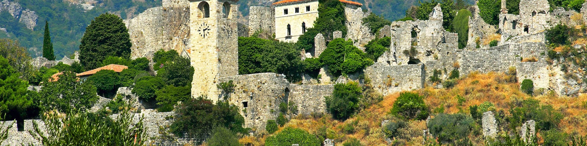 A historic stone fortress with a clock tower and a bell tower, surrounded by trees and rocky terrain.