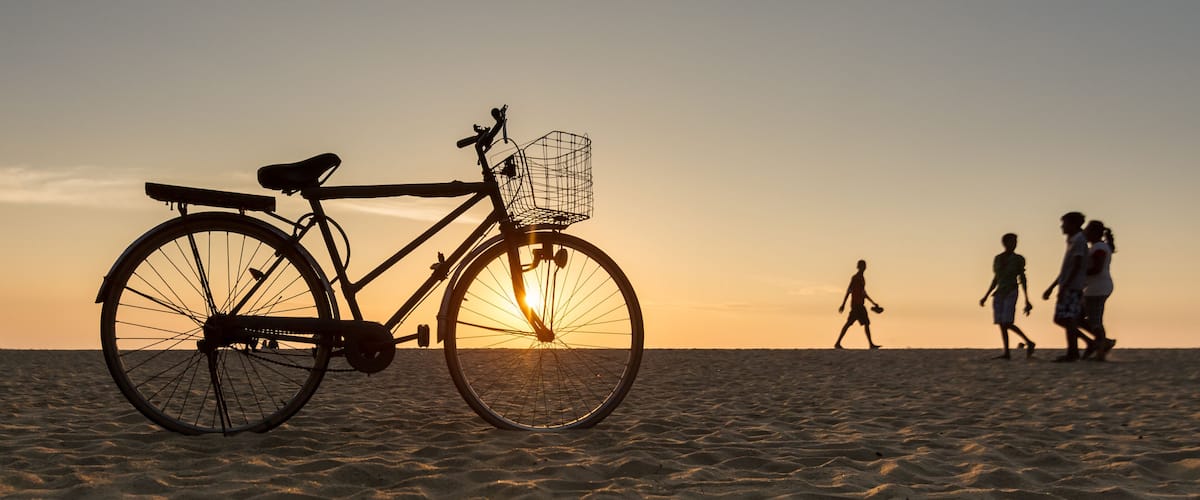 A bicycle on a sandy beach with people walking in the background during sunset.