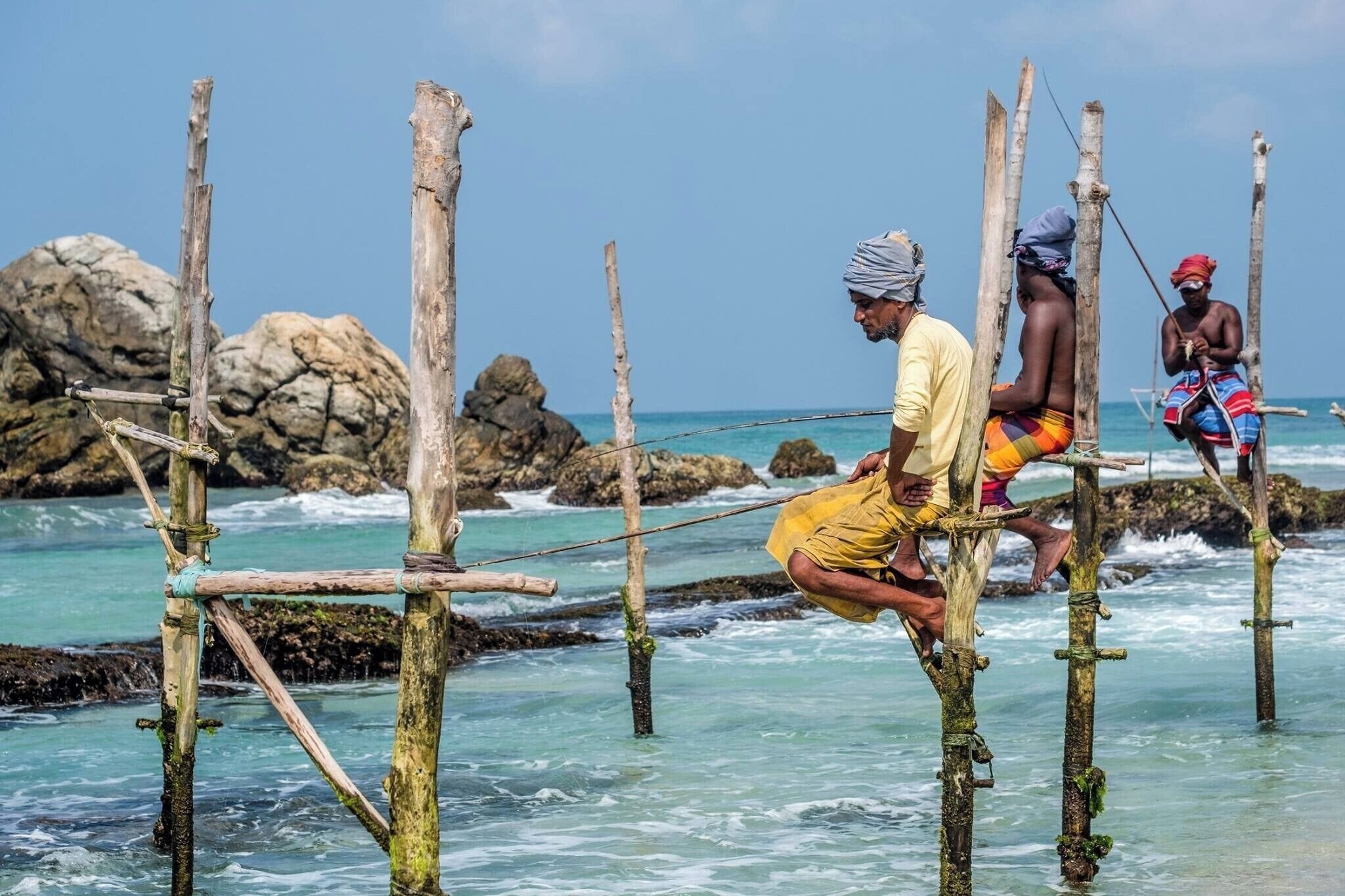 People fishing on wooden stilts in the ocean with rocky formations in the background.