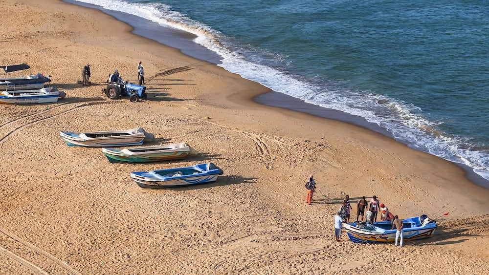 A beach with several small boats lined up along the shore, a person in a blue shirt standing near the water, and a sandy beach with gentle waves.
