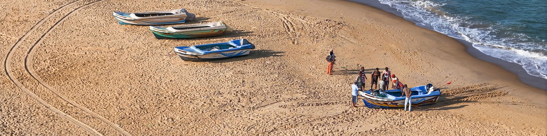 A beach with several small boats lined up along the shore, a person in a blue shirt standing near the water, and a sandy beach with gentle waves.
