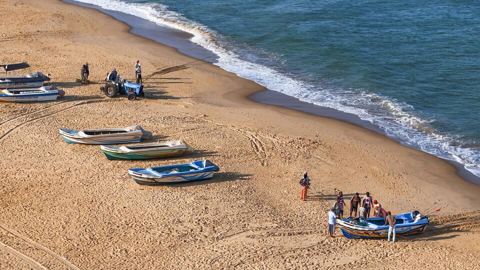 A sandy beach with several small boats lined up, a person on a quad bike, and a few individuals standing by the water's edge.
