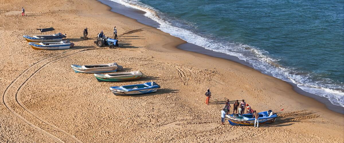 A sandy beach with several small boats lined up, a person on a quad bike, and a few individuals standing by the water's edge.