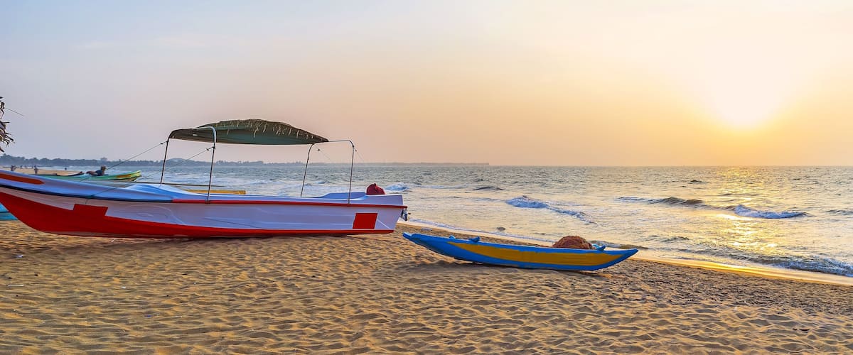 A beach with two boats, one red and white and one blue and yellow, and the sun setting over the ocean.