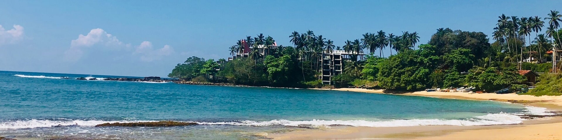 A sandy beach with clear blue water, palm trees, and buildings in the background.