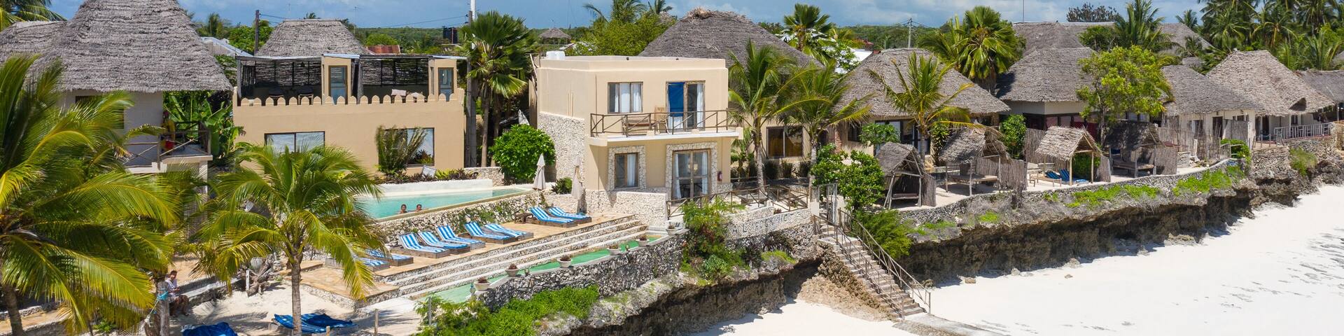 A beach resort with thatched-roof huts, a swimming pool, and palm trees.