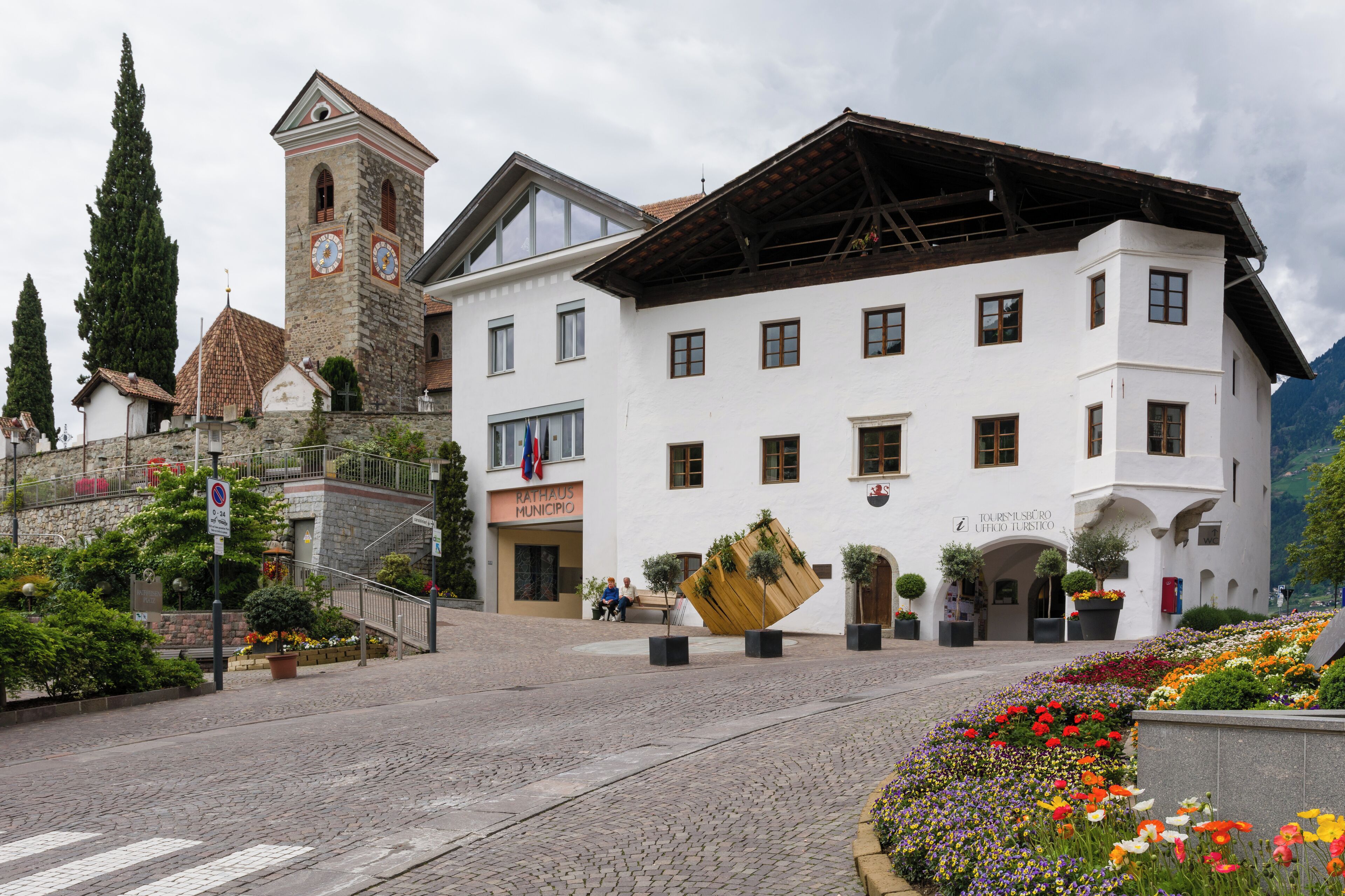 A town square with a church, a white building with a sign reading "RATHAUS MUNIPIO," and a large tilted sculpture.