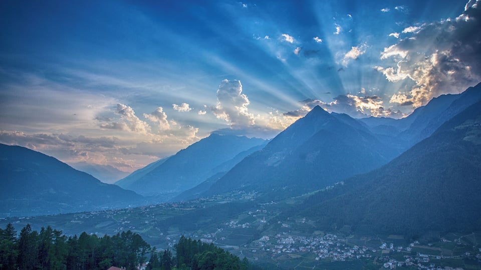 A mountainous landscape with a village at the base, surrounded by lush greenery and a clear sky.