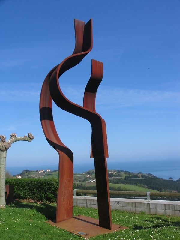 A metal sculpture with a wavy design, set against a backdrop of a clear blue sky and a coastal landscape.