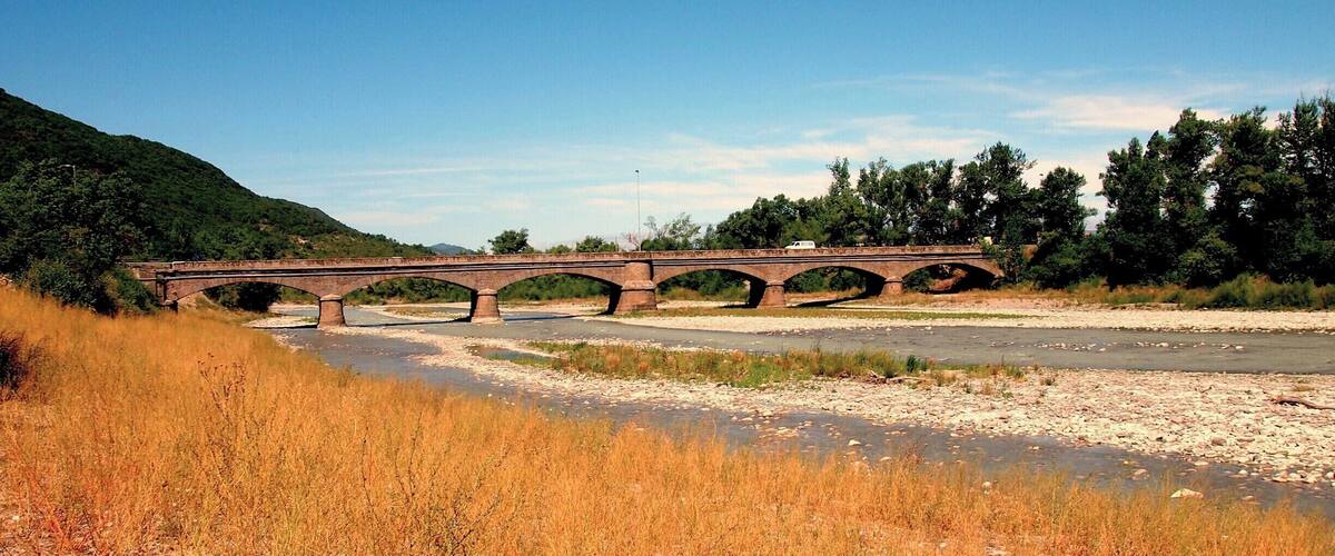 A stone bridge over a river with dry grass in the foreground and trees in the background.
