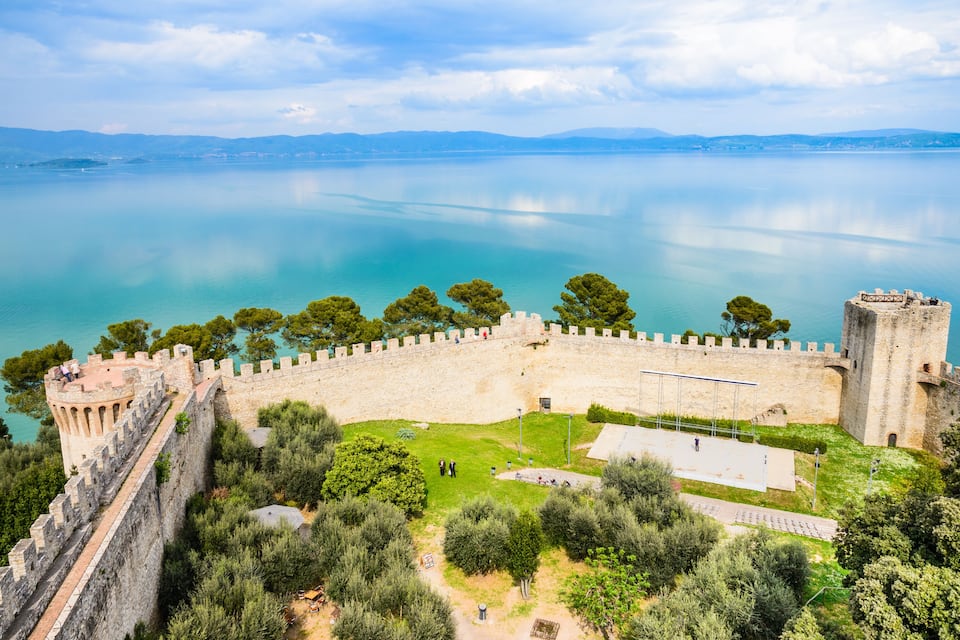 A historic castle overlooking a calm lake with surrounding greenery and distant mountains.