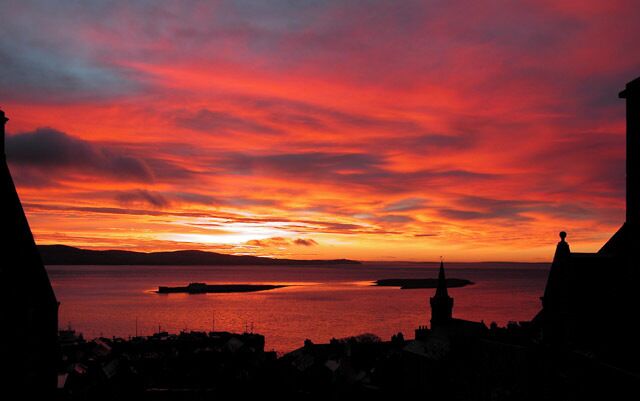 A sunset over a body of water with silhouettes of buildings and a tower.