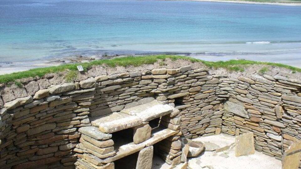 A stone structure by the sea with a view of the ocean and distant hills.
