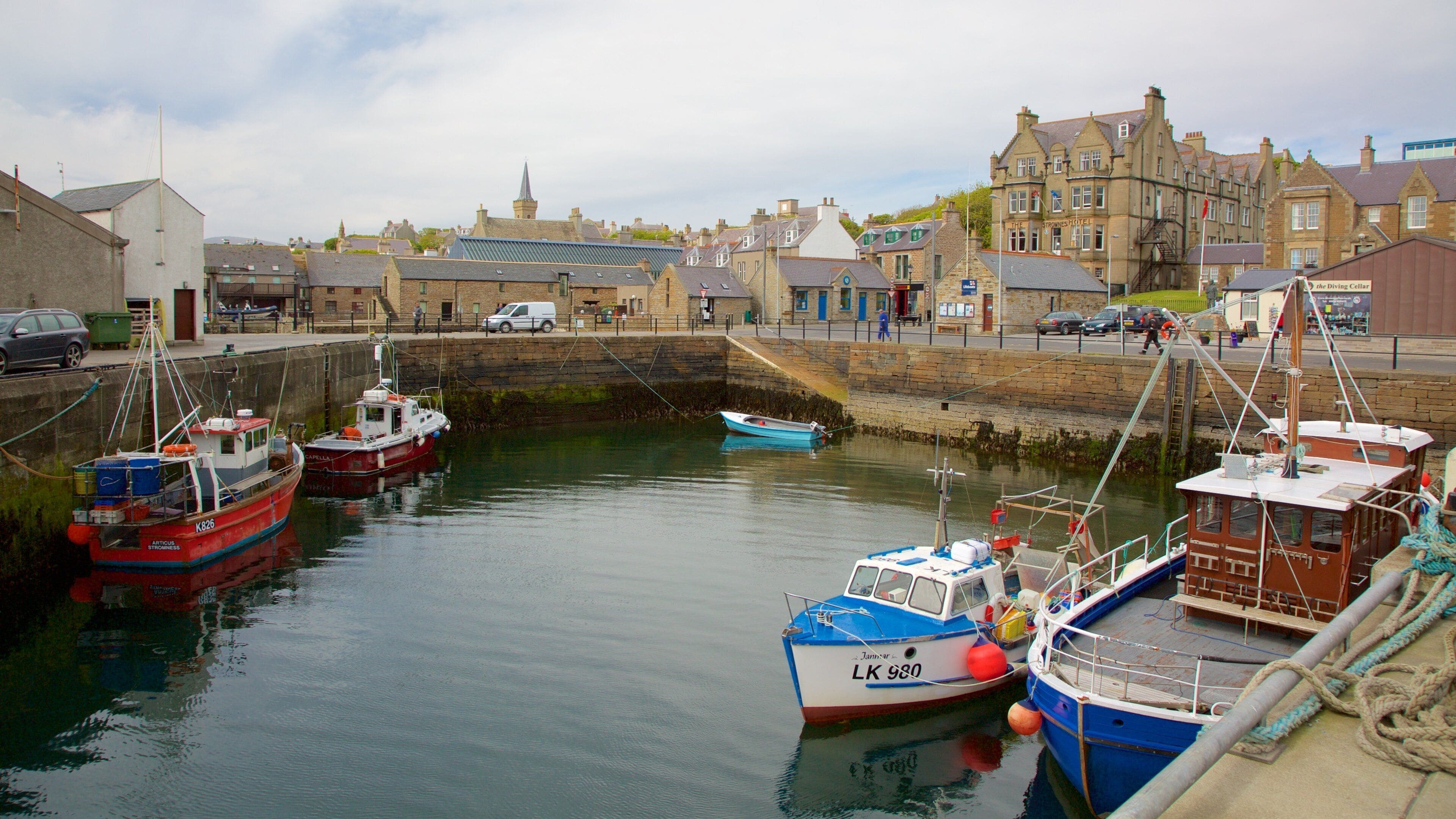 A harbor with several boats docked, a stone wall, and buildings in the background.