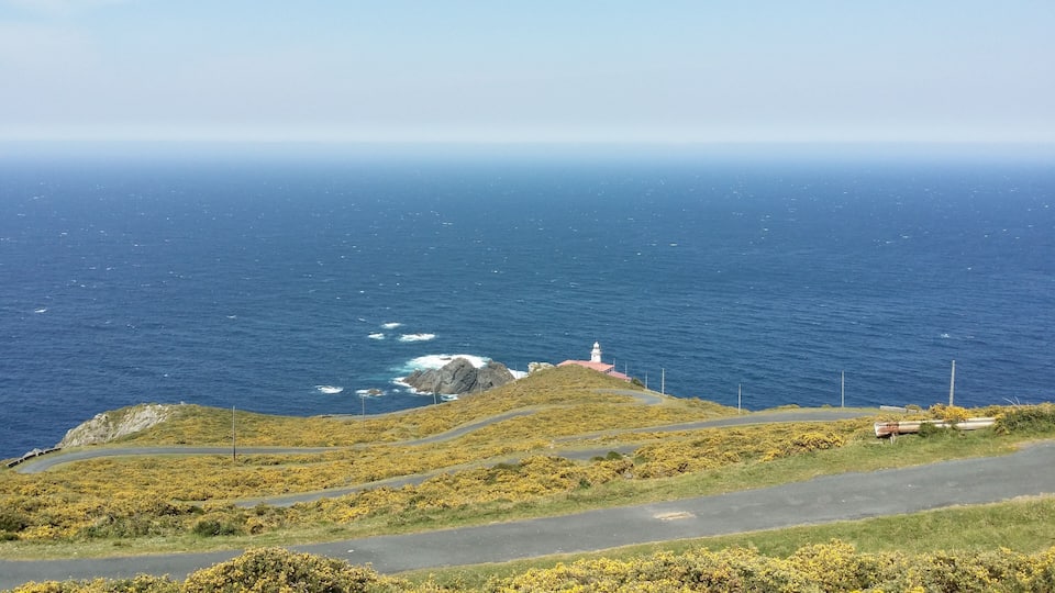 A coastal landscape with a lighthouse on a hill, a winding road, and the ocean.