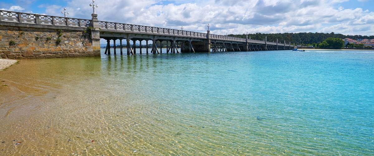 A stone bridge over a clear blue body of water with a beach on one side and a forested hill on the other.