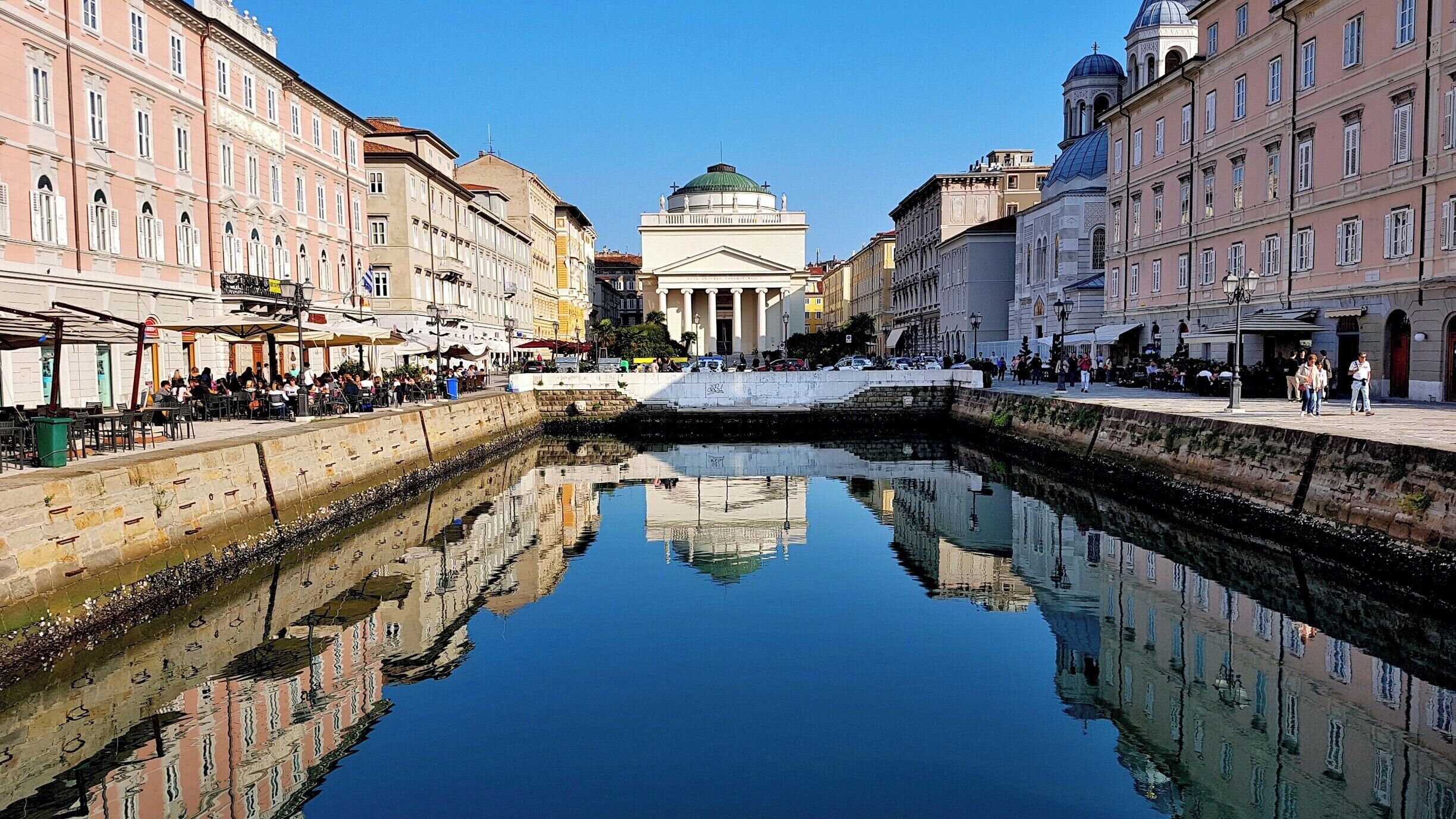 A canal with buildings on either side, a central dome, and people walking along the edges.