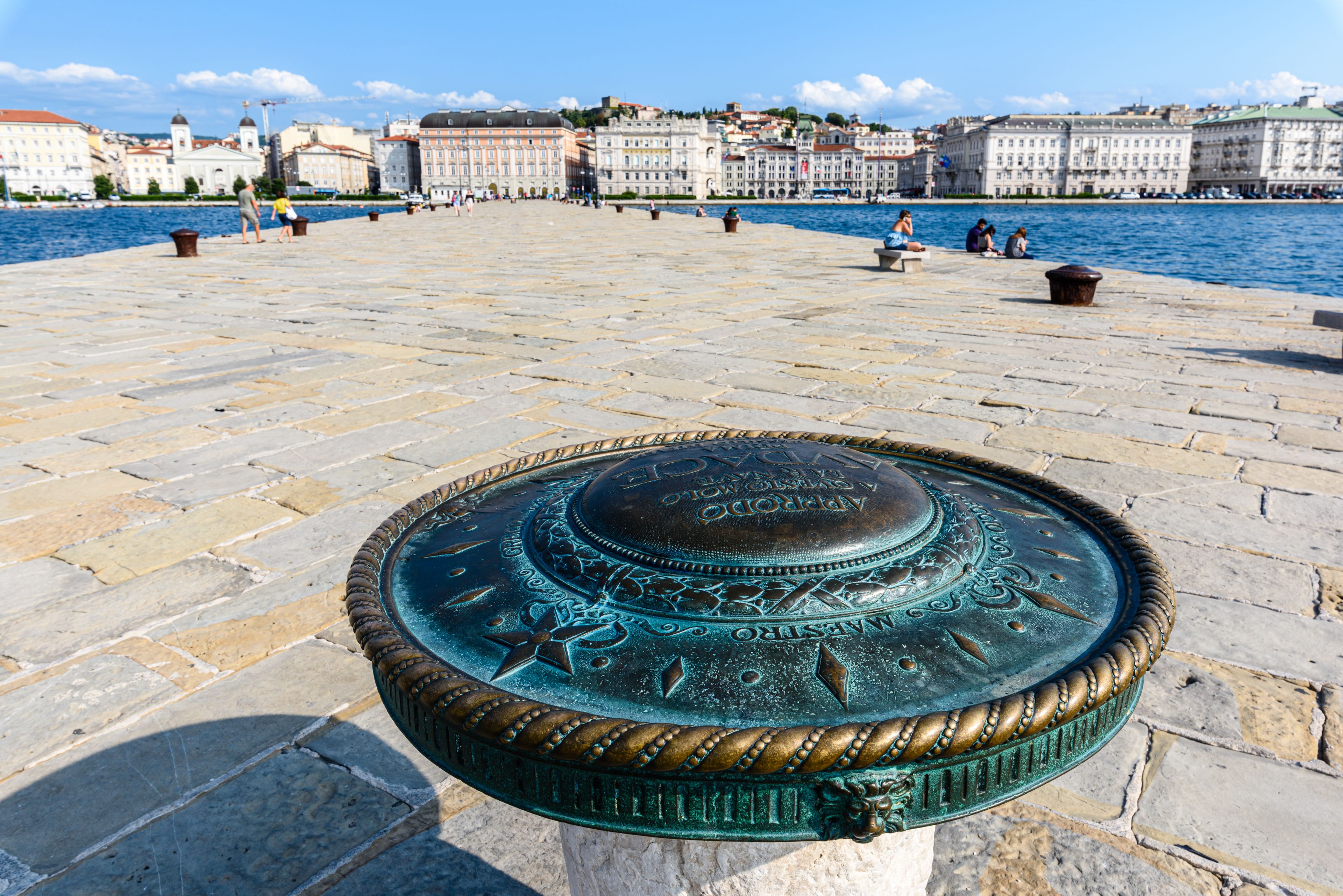 Un paseo marítimo con una fuente de metal decorativa en primer plano y edificios históricos al fondo.