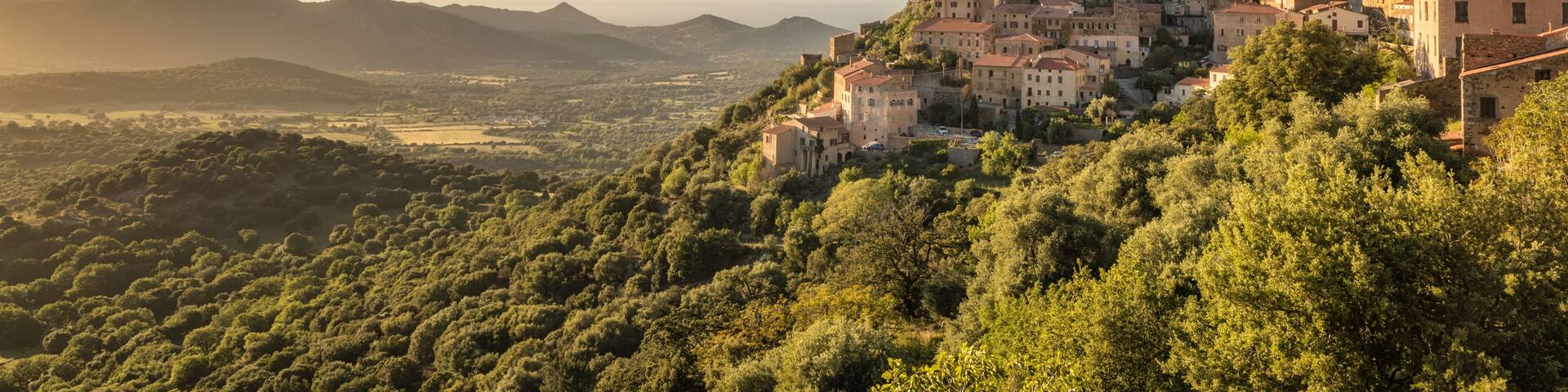 A mountainous town with buildings perched on a hillside, surrounded by dense greenery and a clear sky.