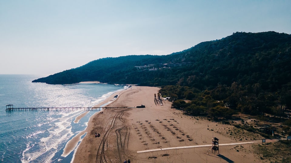 A coastal landscape with a sandy beach, a pier extending into the sea, and a forested hill in the background.