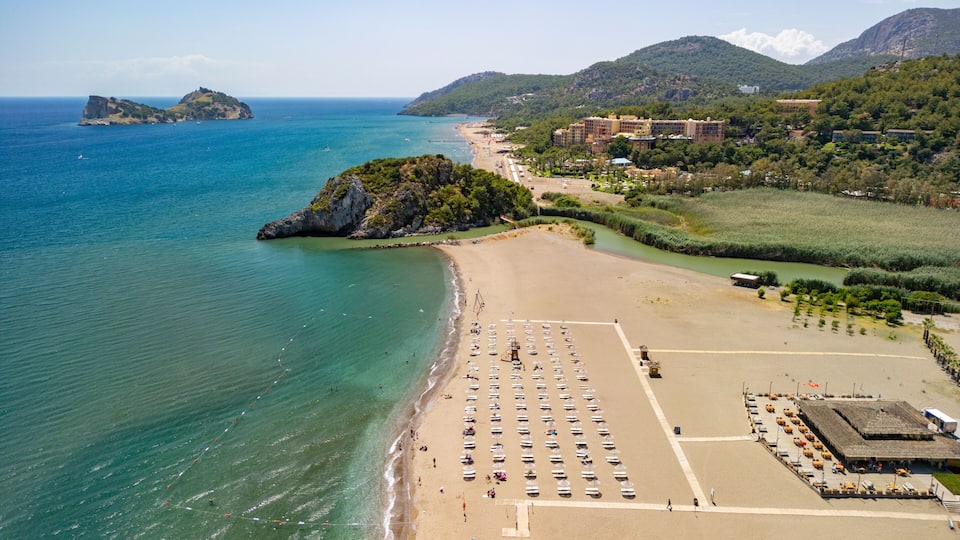 A coastal beach with a parking lot, a small island, and a mountainous landscape in the background.