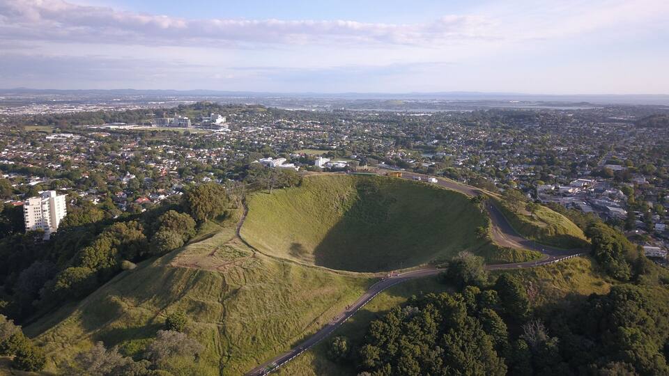 Vista aérea de una ciudad con un parque verde extenso, una carretera sinuosa y montañas distantes.
