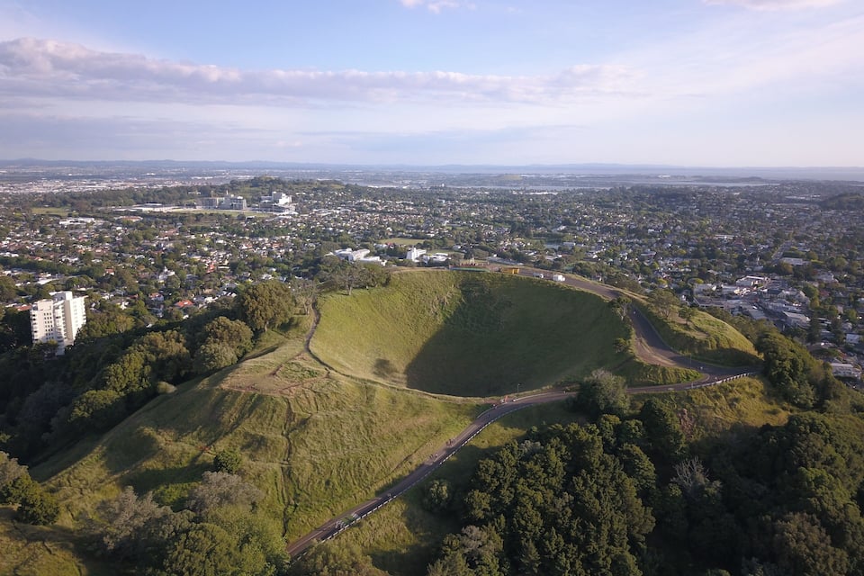 Une vue aérienne d’une ville avec un grand parc verdoyant, une route sinueuse et des montagnes à l’horizon.