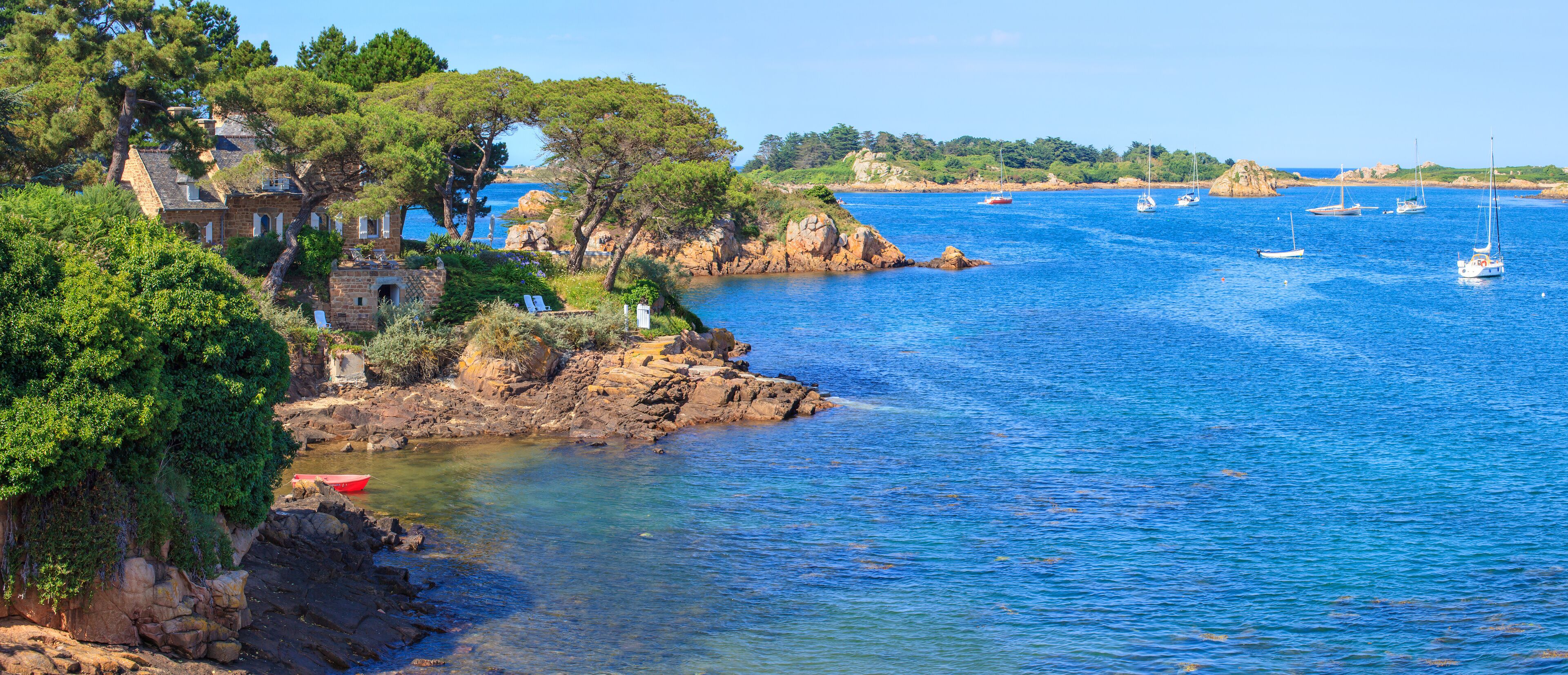 A coastal landscape with a stone house, rocky shoreline, clear blue sea, and several sailboats.