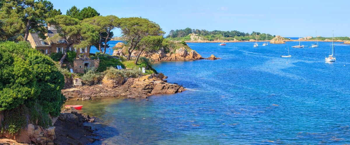 A coastal landscape with a stone house, rocky shoreline, clear blue sea, and several sailboats.