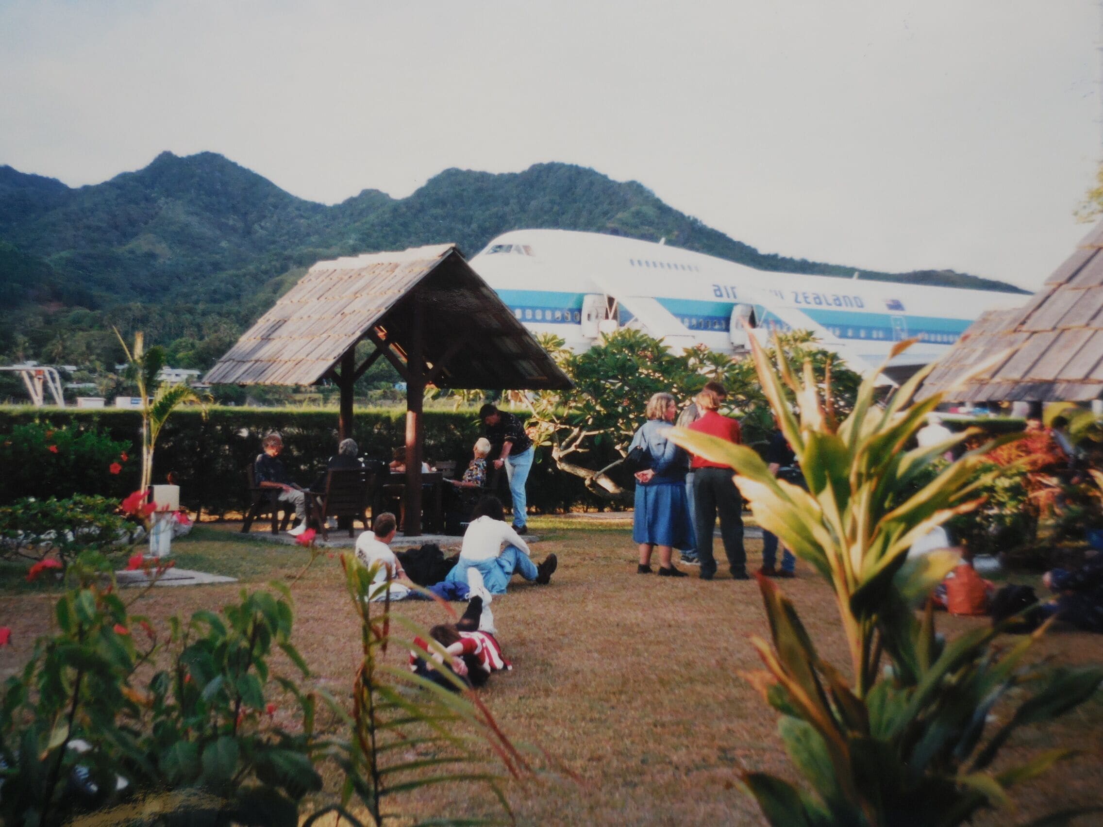 A group of people gathered around a wooden structure in a garden with a large airplane in the background.